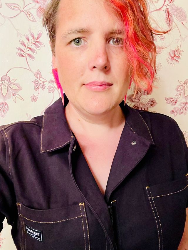 Miriam in black coveralls with asymmetrical hair that includes orange streaks, and large neon lighting bolt earrings.

