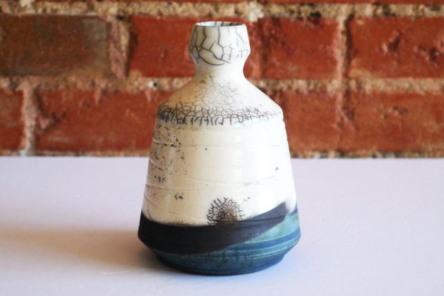 A bottle with a flared base tinged blue-green from oxidized copper, a black-then-white swoop of bare clay on walls that slope in towards the neck, and then glossy white crackle glaze. The cracks have only blackened in very specific places – a circle near the base, a sliver along the shoulder, and around the head and rim of the bottle itself.

