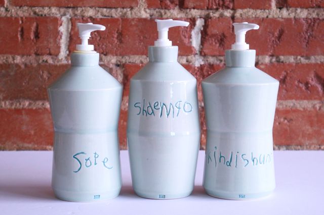 Three angular light blue bottles in a line, with white pump dispensers in each. The outer two angle in towards the middle, the center one angling out, so they fit together in order. A child's misspelled handwriting is carved in and glazed darker blue: sope, shaempo, and kindishunr.
