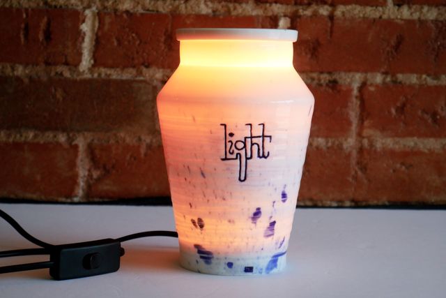 An urn-like jar with a power cable glowing amber from inside. The shape is angular - out from a narrow foot to a wide shoulder, and then back in to an upright rim with a slab lid over the top. The side says light in a hand-carved upright slab cursive with connected letters, and there are splashes of blue.
