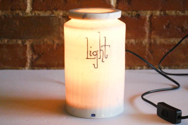 Tall cylindrical porcelain jar glowing warmly from the inside. The word light is handwritten on the side with a blue inlay and playful slab serif lettering.
