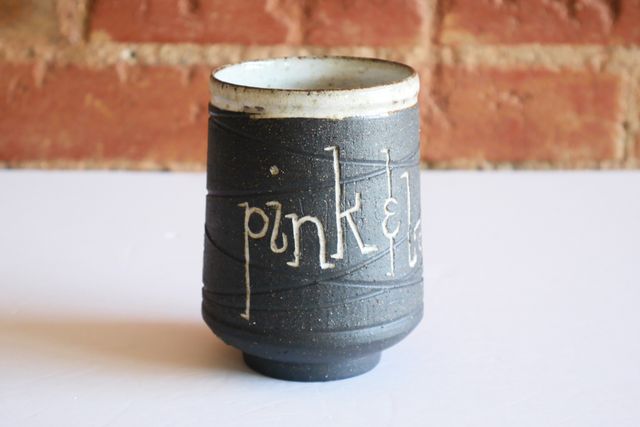 Ceramic tumbler with an inset foot ring, supporting a rounded base and angled walls sloping towards the blue-glazed rim. Most of the piece is black ceramic with a grooved texture, and inlaid white text that says pink & lavender & blue.
