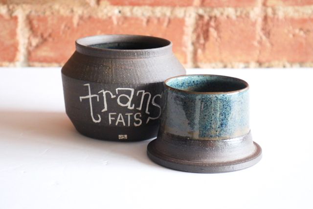 An open butter bell in two pieces, the upright jar and overturned lid revealing a bell inside – now upright. On the side of the jar it says trans fats in a hand-carved slab serif inlaid with white.
