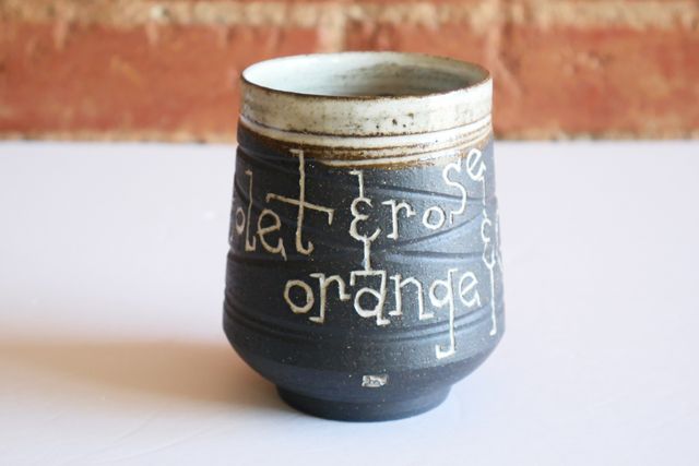 Ceramic tumbler with an inset foot ring, supporting a rounded base and angled walls sloping towards the blue-glazed rim. Most of the piece is black ceramic with a grooved texture, and inlaid white text that says orange & coral & white & violet & rose.
