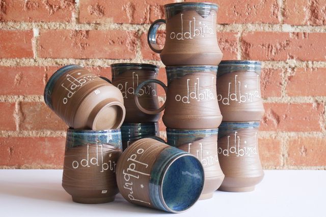 A stack of mugs that all say OddBird in playful hand-carved lettering. The mugs and tumblers all have a deep foot, and rounded base. The tumblers slope in towards the lip, while the mugs have a slightly concave curve in, and a round handle.
