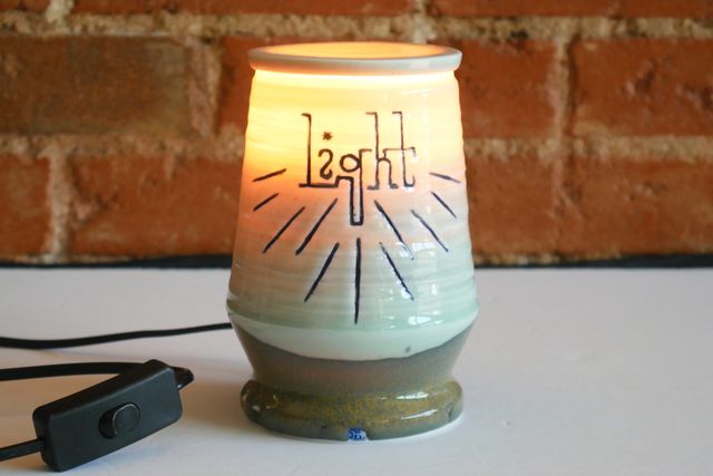 A blue jar with green-and-gold base, but the top half and lid are glowing amber from inside. The word light is carved near the top in a sort of handwritten slab serif, with blue rays emanating from the text.
