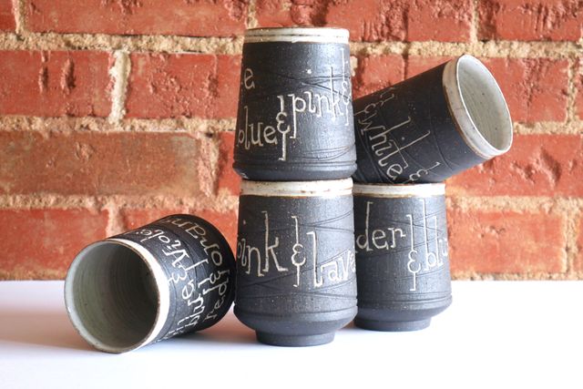Stack of black tumblers with white glaze inside and on the rim, and hand-carved lettering inlaid white listing the colors of various pride flags. Blue & pink & white & pink & blue, for example, the colors of the trans flag.
