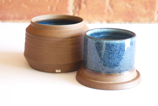 A butter bell sitting open, with raw brown clay on the outside, but a glossy speckled floating blue glaze covering all the interior surfaces of the cylindrical bell and wide jar.
