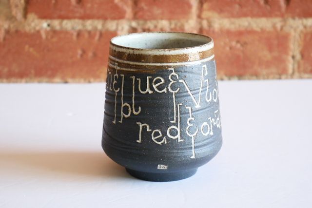 Ceramic tumbler with an inset foot ring, supporting a rounded base and angled walls sloping towards the blue-glazed rim. Most of the piece is black ceramic with a grooved texture, and inlaid white text that says red & orange & yellow & green & blue & violet.
