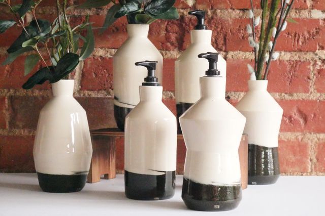 Six ceramic bottles, each with a black clay base that transitions with a hard edge and slight swirl to white clay in the first several inches. Some have flowers, other have pump dispenser corks for something like soap or lotion. Several are simple cylinders with an angular shoulder and neck, but a couple have an extra angular waist either pulled in like an hourglass or pushed out like a diamond.
