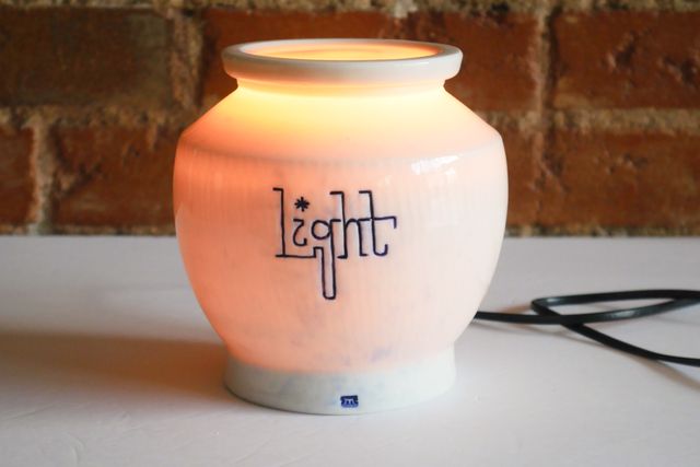 white porcelain jar glowing amber from the inside. From the base it flares out and up like a honey pot, and then angles back in near the top, to an opening that matches the base. The side is textured in vertical chatter, and light is carved in an upright sort of slab cursive. The lid is on and also glowing.
