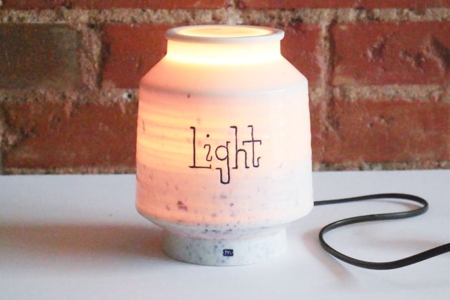 A porcelain jar that's glowing amber from the inside, and has the word light hand-carved on the front in a sort of slab serif font. The lid is on and also glowing warmly. The jar has a lantern-like shape, narrow cylinder at the base and rim, then angling out to a wider cylinder for most of the body. There's a texture carved around the wide section, so that it glows in subtle rings.
