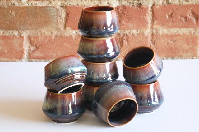 Nine small ceramic cup stacked and displayed in different orientations. They angle out at the base, and then back in towards the rim.
