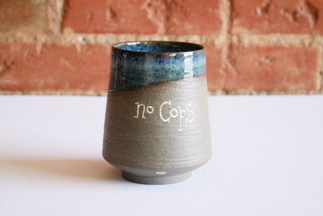 Ceramic tumbler with an inset foot ring, supporting a rounded base and angled walls that gently slope towards the blue-glazed rim. Most of the piece is exposed toasty gray ceramic with a grooved texture, and inlaid white text that says no cops, no cages, no borders.
