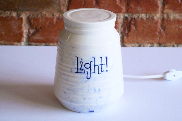 Angular white porcelain jar that angles out at the base, and then gently back in towards the top, with a small shoulder to match the bass, and an upright rim holding the lid. There's a white lamp cord coming from the back with a small white switch.
