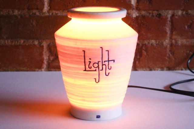 Tall porcelain jar with a narrow base and wide angular shoulder, glowing warmly from the inside. The word light is handwritten on the side with a blue inlay and playful slab serif lettering.
