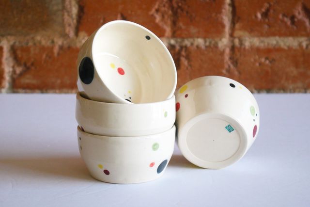 Four small bowls with a flat base and linear walls that angle out then up -- in white clay with colorful floating dots spread out in different sizes.

