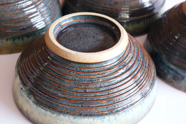 The underside of a bowl, with a deep inset foot-ring -- glazed with the same deep red-brown and floating blues. The foot angles out from a flat ring in toasty bare clay with dark speckles, then cuts back in diamond-like towards the outer walls. The outside of the bowl is textured with deep grooves around with red-brown peaks, and deep blue valleys. The texture ends abruptly about an inch from the rim, where the whiter glaze reveals a chattering texture over an otherwise flat ribbon-like lip.
