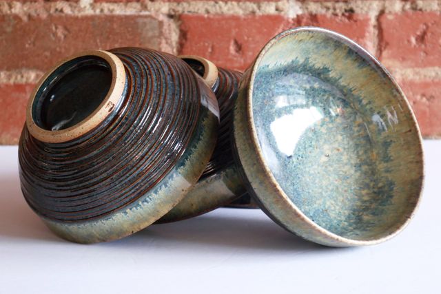 Three bowls stacked as though drying, one lip-down, and the others leaning with their rims against the foot of the previous -- showing the glazed base inside the foot-rings, and the textured sides. The fourth again is leaning bowl-out, to show off the inside form and nebula-like splash of glazes.
