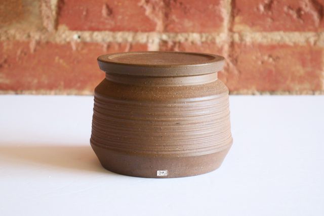 Closed, with the bell lid hidden away, it appears to be a brown clay jar that angles out from the base, and then slopes in gently with a texture of grooved rings. Near the top it angles in more dramatically to a short vertical lip.
