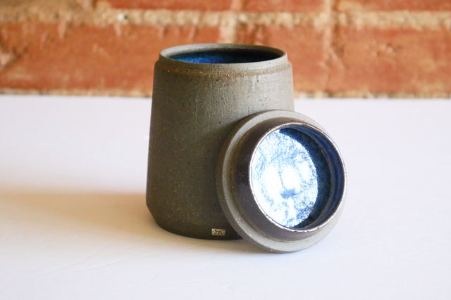 Small jar with the lid leaning against the outside -- revealing a hint of bright speckled blue inside a gray textured and refined but unassuming jar.
