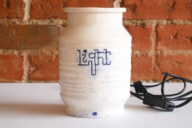 The light is off now, revealing the piece is a bright glossy white, with blue text, a blue makers mark, and a splash of faded blue across the front. The text is strung together, almost as though written in string. There's a black cable coiled beside the jar with a rocker switch.
