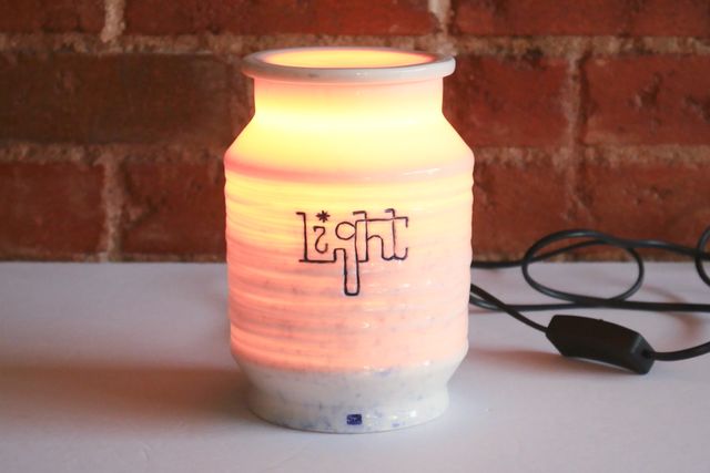 White porcelain jar glowing a warm amber from within. It's shaped a bit like a paper lantern, a wide textured cylinder that angles in near the rim and base which become vertical cylinders again at a narrower width. The word light is carved and inlaid with blue wash high up on the main body, and there's a thin glowing lid on top.
