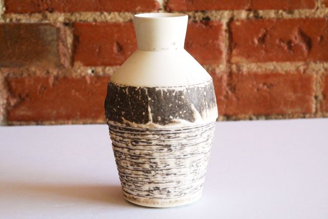 At the shoulder, there's a darker vertical section where the texture changes from carved grooves around, to organic ridges like plaster that's been spread but not smoothed - and then a line where the form angles in towards the neck, bare white clay with occasional spray of color.